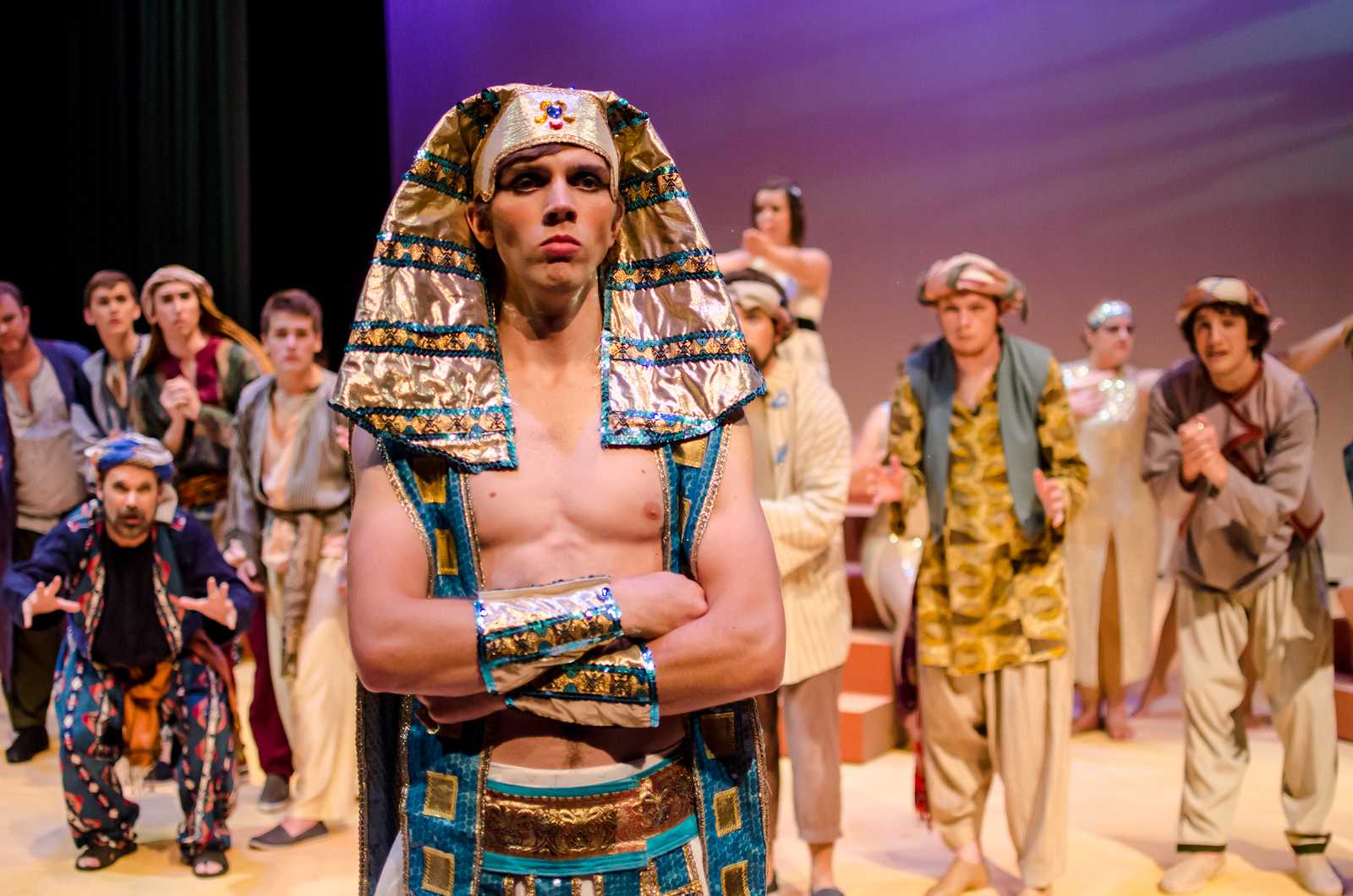 Joseph, a young man wearing the costume of an Egyptian pharoah, crosses his arms and stares out towards the camera with a determined expression. A group of men who are mostly his brothers, all wearing Egyptian clothing, are arrayed in pleading positions behind him.